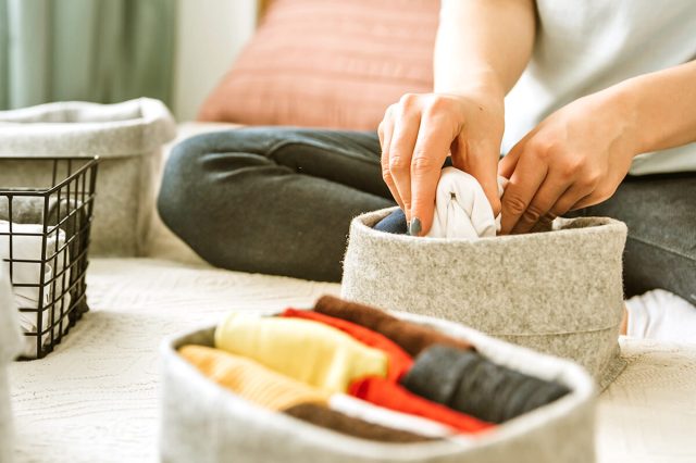 Anonymous woman folding small clothing items into a circular, gray felt storage bin. In front of her are other storage bins in various shapes and materials.