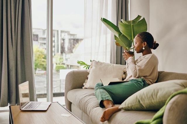 Woman sitting on a beige couch drinking a cup of coffee. Her apartment is nicely decorated with plants and cozy textures and a laptop is sitting on the coffee table in front of her.