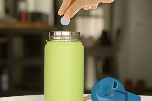 Close-up of a person's hands dropping a denture tablet into a lime green insulated water bottle. The water bottle lid is next to it on the table.