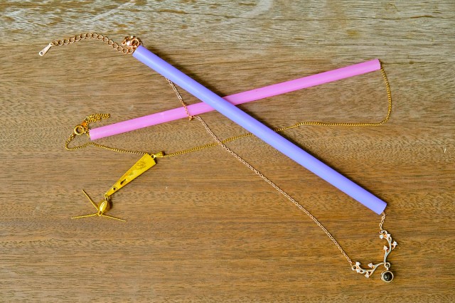 Two chainlink necklaces (one silver, one gold, both with pendants) looped through plastic straws to keep them from tangling. The straws are overlapping, creating an 'X' shape on a wooden table.