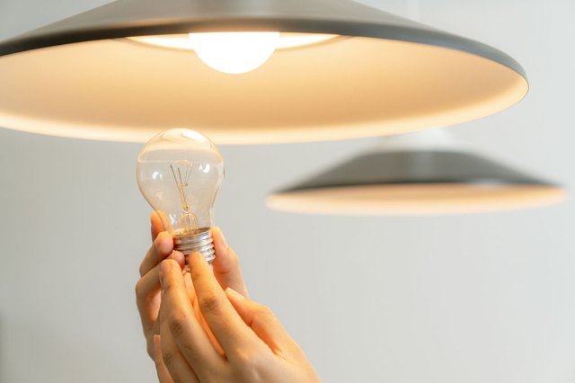 A close-up shot of a person's hands holding a new lightbulb up to a farmhouse-style pendant light that is turned on and features a different style of lightbulb.