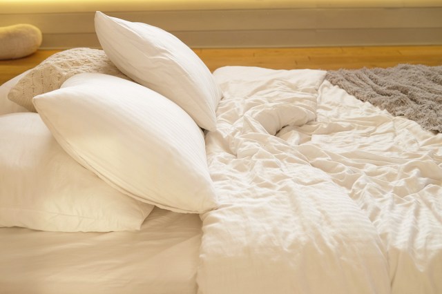 A close-up of a bed with lots of pillows, a fluffy duvet with a white cover, and a fuzzy gray throw blanket at the foot of the bed.