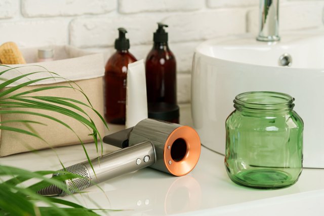 A hair dryer on a bathroom countertop next to a green glass jar and two amber-colored glass soap bottles with pumps.