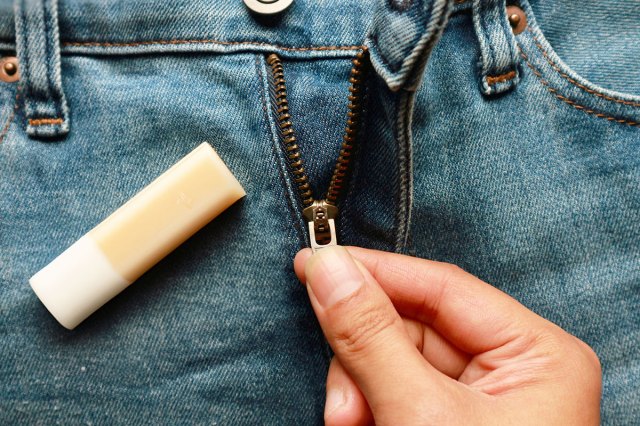 Anonymous person pulling down a zipper on a pair of jeans. The jeans are laid flat on a surface. Placed on the jeans beside the zipper is a tube of lip balm.