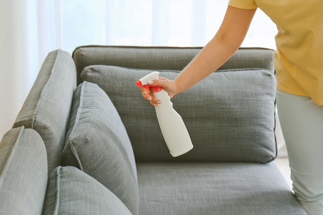 Anonymous woman wearing a yellow shirt and light gray pants holding a spray bottle up to a medium-gray sectional as if to spray the upholstery.