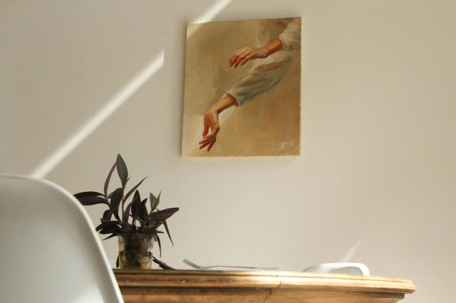 Frameless oil painting hung on a wall depicting a person's hands. On a wooden table below is a fork and a colorful plant. Two white resin chairs are tucked in around the round table.