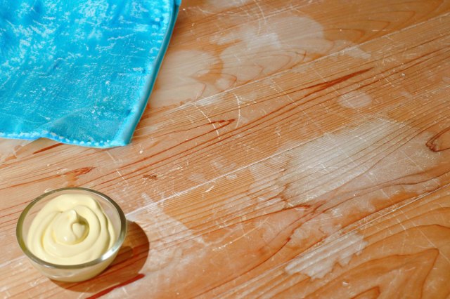 A close-up of a wood table with lots of scratches and water stains. There is a glass bowl of mayonnaise on the table, as well as a blue cleaning cloth.