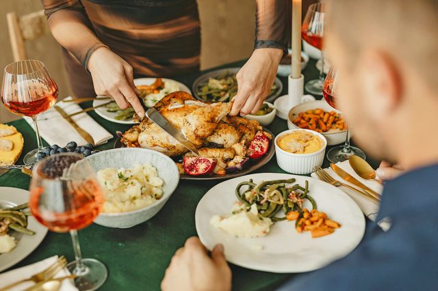 Two anonymous people at a Thanksgiving dinner table. One person is cutting into the turkey while the other is waiting patiently and has a plate filled with green beans, mashed potatoes, and carrots.