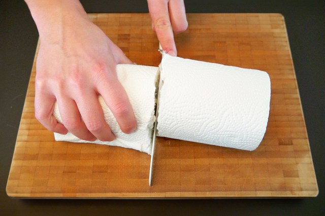 Aerial view of a person cutting a paper towel roll in half with a knife on a wooden cutting board.