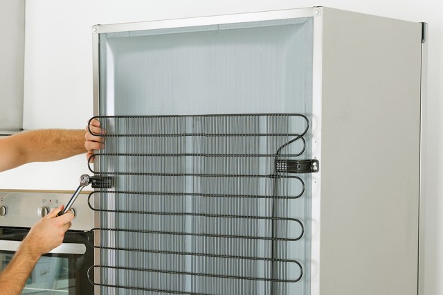 An anonymous person tightening a part on the back of a refrigerator that keeps the coils attached. The refrigerator has very clean coils and is in a kitchen.