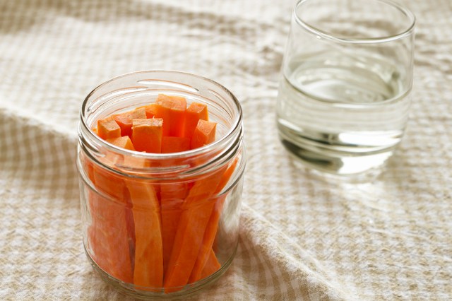 Matchstick-cut carrots sitting in a Mason jar on a flat surface covered with a gingham tablecloth. Next to the carrots is a glass of water.