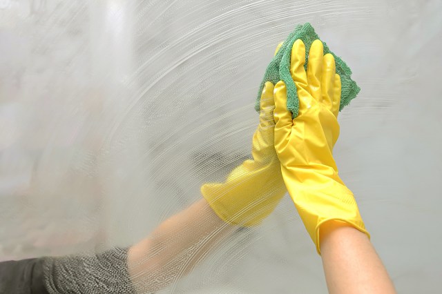 Anonymous person wearing a yellow cleaning glove wiping a large bathroom mirror with a green microfiber cloth.