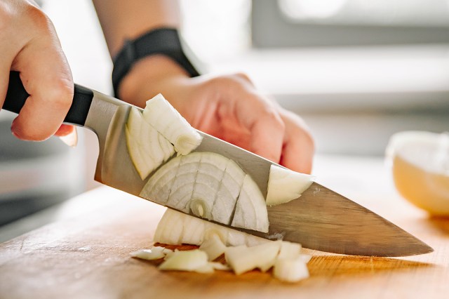 Anonymous person dicing a white onion on a wooden cutting. board. The person is using a chef's knife and wearing a smart watch.