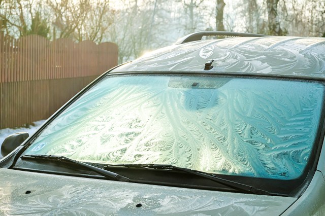 A close-up shot of an icy car windshield on a winter morning. There are bare trees and snow in the background.