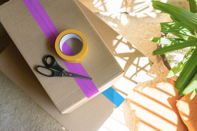 A cardboard box sealed shut with purple masking tape. A roll of yellow masking tape and a pair of scissors are sitting on top of the box.