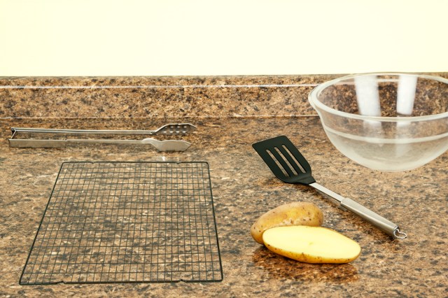 Black metal cooling rack with a grid pattern on a countertop next to a potato, spatula, and clear glass bowl.