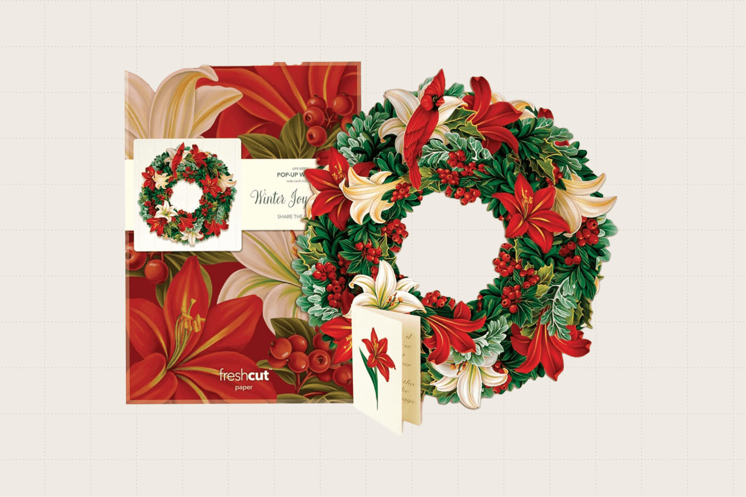 The FreshCut Paper pop up holiday wreath, measuring 15 inches in diameter. It features faux holly, red and white lilies, a cardinal, and evergreens.