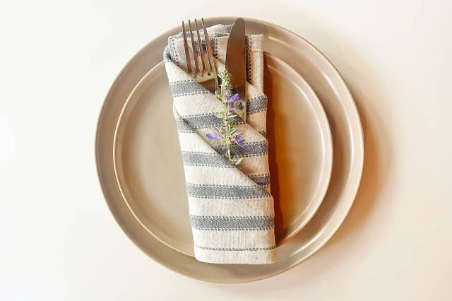 A folded dinner napkin with three pockets holding a fork, knife, and a sprig of lavender sitting on a salad plate that's on top of a dinner plate on a white surface.