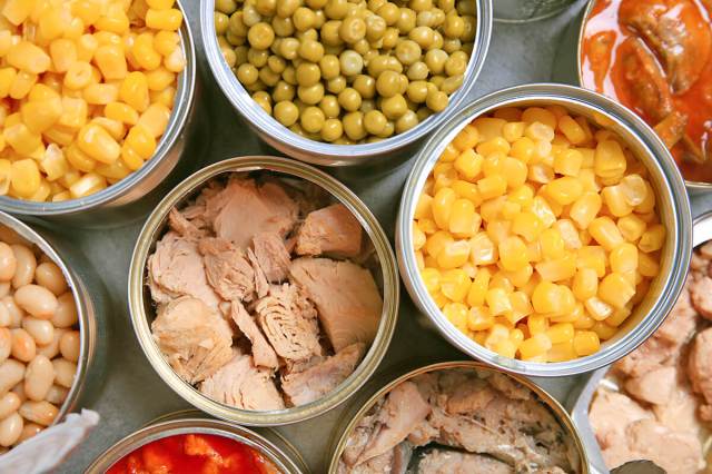 Multiple open cans of food viewed from above including tuna, corn, peas, and white beans.