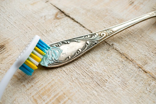 A toothbrush with toothpaste on it scrubbing the engraved handle of a silver utensil.