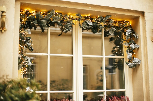 A garland inside a doorframe on the outside of a tan house. The garland has frosted leaves and twinkle lights.