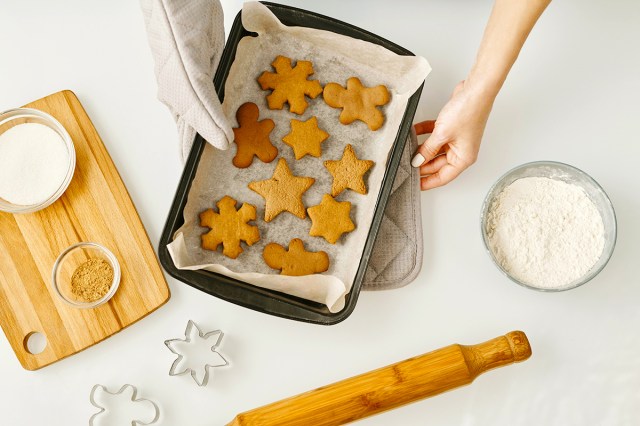 Anonymous person holding a baking sheet lined with parchment paper that has gingerbread cookies on it in the shape of snowflakes, stars, and gingerbread men. On the table, there's a bowl of flour, a rolling pin, a bowl of sugar, and a cutting board.