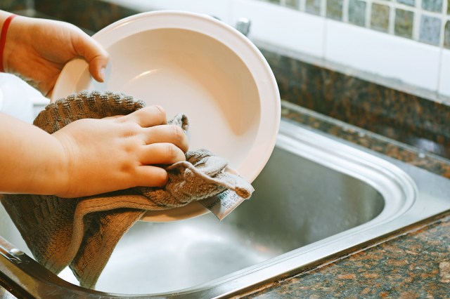 An anonymous person standing at the kitchen sink scrubbing a shallow bowl with a cotton tea towel.