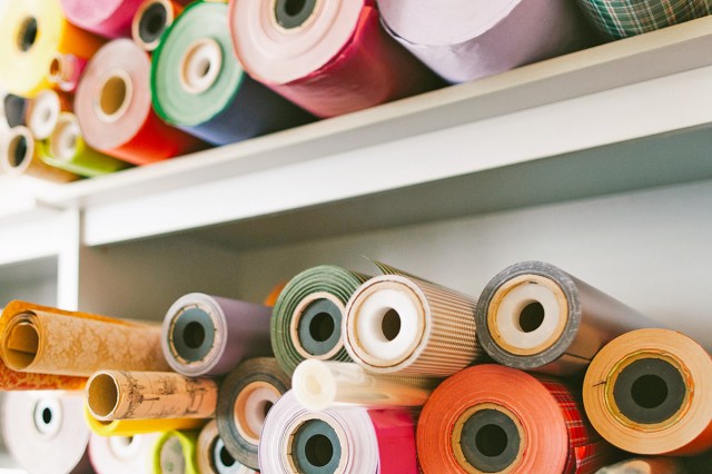 Large rolls of gift wrap, all in different patterns, stacked horizontally on a white bookshelf. Some rolls are in focus, others farther away are not.