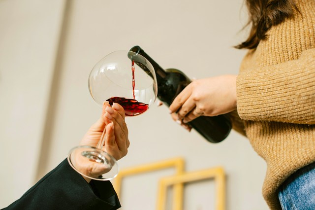 Anonymous woman wearing a mustard color knit sweater and jeans pouring a glass of red wine for another anonymous woman wearing a black coat.