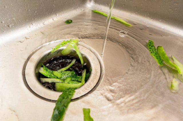 Close-up of a drain leading to a garbage disposal in a stainless steel sink. The water is running and there are vegetable scraps in the drain.