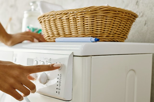 Anonymous person pressing one of the cycle buttons on their laundry machine. On top of the machine is a blue dry erase marker without the cap and a laundry basket.