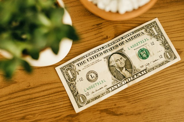 A U.S. dollar bill sitting face-up on a wooden tabletop. Next to it and out of focus is a potted plant and a dish.