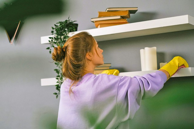 A young woman wearing an oversized purple sweatshirt and yellow cleaning gloves dusting two open shelves on a gray wall. The shelves hold decorative items like books, candles, and plants.