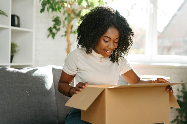 A woman opening a large cardboard box and peering inside. She's sitting on a gray couch in an apartment with lots of natural light and a potted tree.