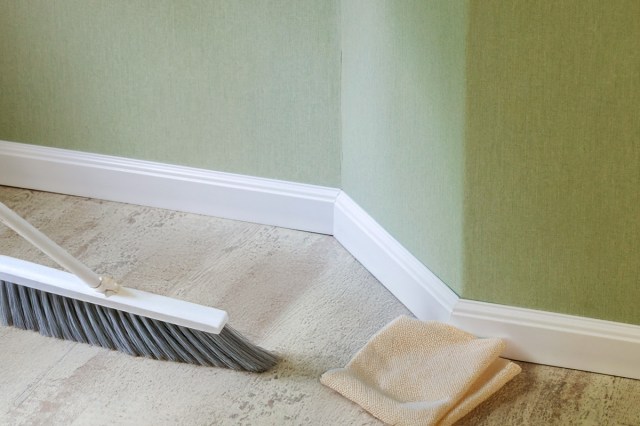 An image of white baseboards against a green wall. On the laminate floor is a household broom and a dry microfiber cloth.