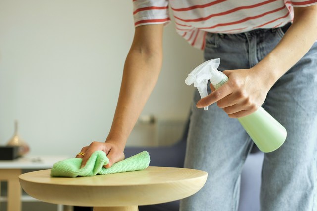 Anonymous woman in a white and red striped short-sleeve shirt wiping down a side table with a spray bottle and a microfiber cloth.