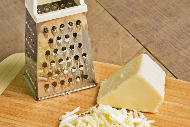 A block of white cheese on a wooden cutting board with some shavings next to it and a box grater.