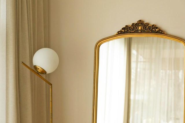 An arched, gold-frame mirror leaning up against a beige wall. Next to it is a modern-style floor lamp.