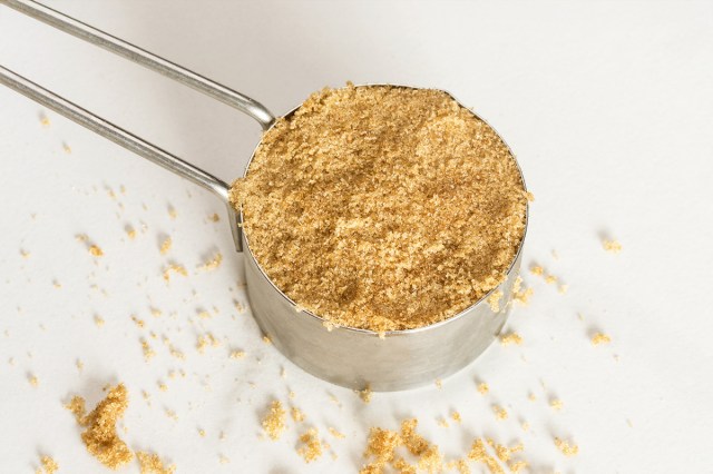 A close-up, overhead shot of a measuring cup full of brown sugar with a little bit sprinkled on the white surface it's sitting on.