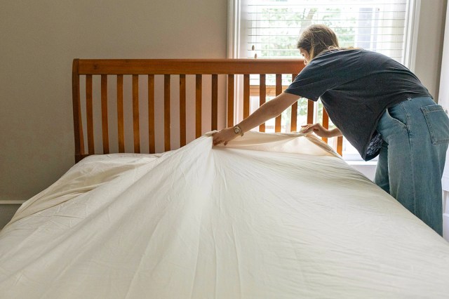 Anonymous person making a bed. They are in the process of adding a top sheet and the bed has a wooden headboard.
