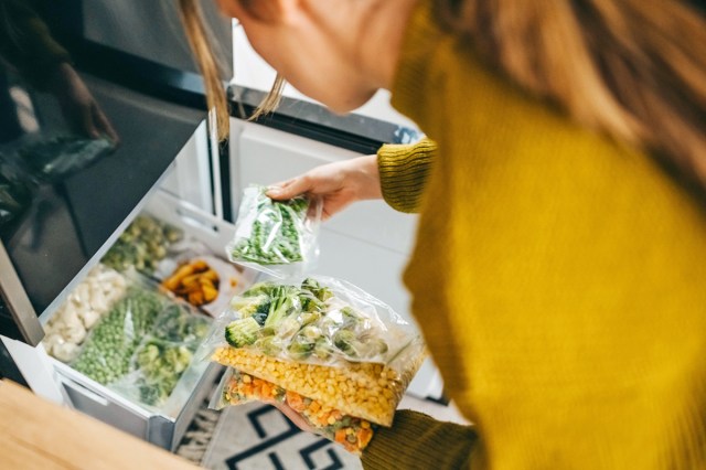 Anonymous woman wearing a mustard-colored shirt going through bags of frozen vegetables in a bottom freezer.
