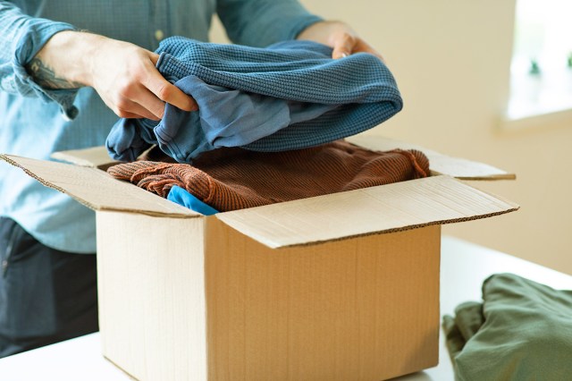 Anonymous person putting folded clothes into an open cardboard box on a table.