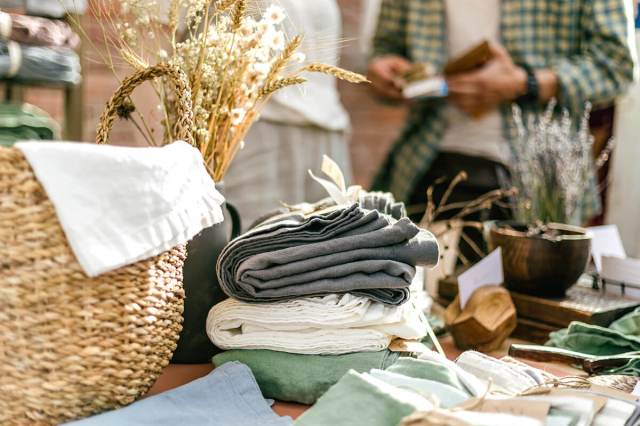 A table at a yard sale stacked with textiles, a basket, and wooden home decor items. Blurred in the background is a man paying a woman with cash.