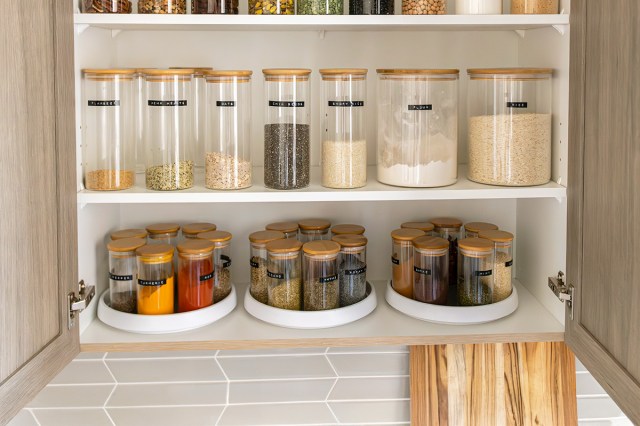 An open kitchen cabinet filled with well-organized spices in labeled, clear jars. Some are arranged on lazy Susans.