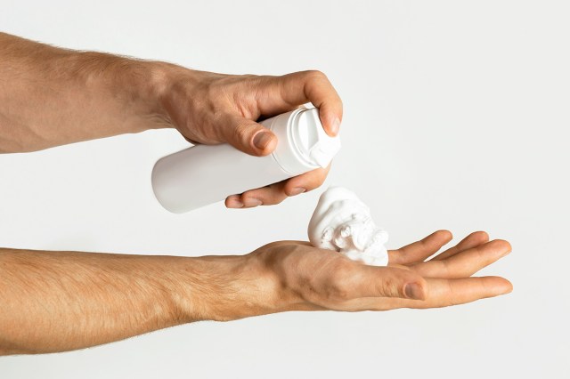 Anonymous person dispensing foamy shaving cream from a white, unlabeled can into their palm.