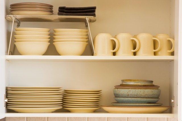 The inside of a well-organized kitchen cabinet, showing plates, bowls, and cups. On the upper shelf, there's a shelf riser to make use of vertical space and allow for more storage.