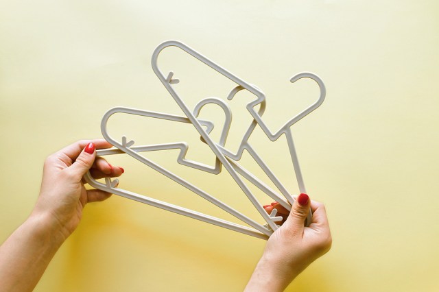 Anonymous person with red nail polish holding three plastic baby hangers against a yellow backdrop.
