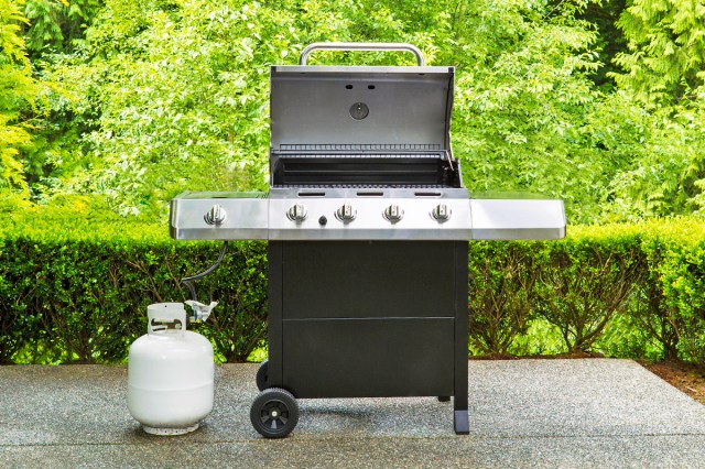A gas grill with the lid open that's hooked up to a propane tank. The grill is on a concrete surface and there are shrubs and trees in the verdant background.