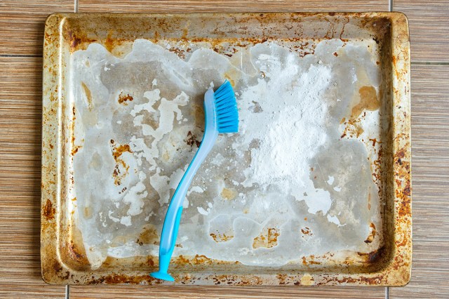 A heavily stained sheet pan with a dish brush on top of it and some baking soda sprinkled on the surface.
