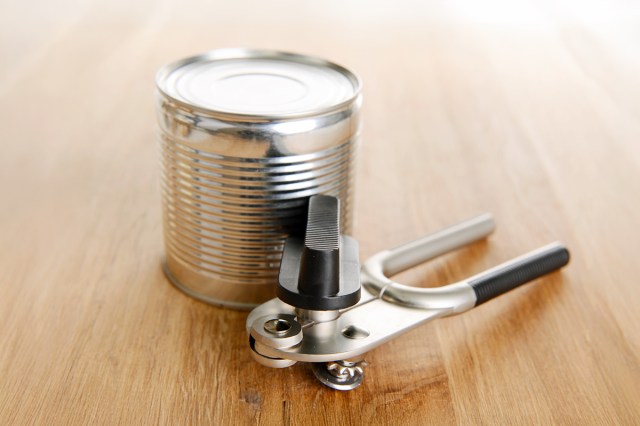 An unlabeled aluminum can on a wooden surface next to a metal and black can opener.